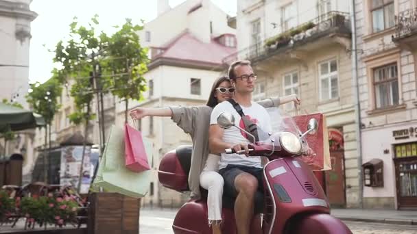 Adorable joven feliz en gafas de sol con estilo con compras a caballo en la moto con su novio a través de la calle urbana — Vídeo de stock