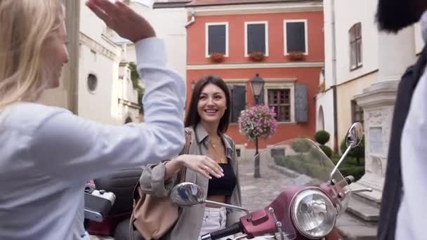 Preciosa mujer alegre sentada en el scooter y dando cinco a sus amigos de razas mixtas en el patio de la ciudad con hermosos edificios — Vídeos de Stock