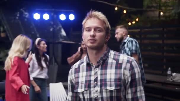 Snygg upprymd blond kille i rutig skjorta dansa under inflammatorisk musik med sina vänner i trädgården huset på kvällen — Stockvideo