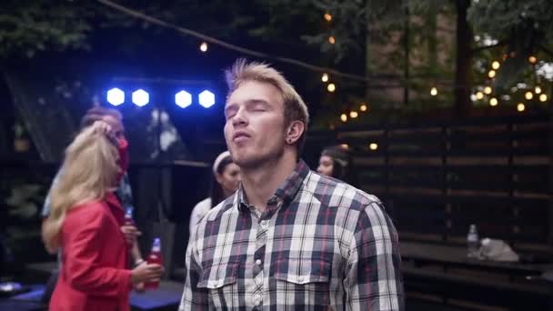 Knappe 30s blonde man in stijlvolle shirt zorgvuldig dansen in de buurt van zijn vrienden in de tuinhuis in de avond — Stockvideo