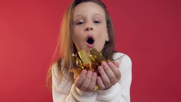 Menina adolescente muito jovial com cabelo longo soprando confete dourado no fundo vermelho — Vídeo de Stock