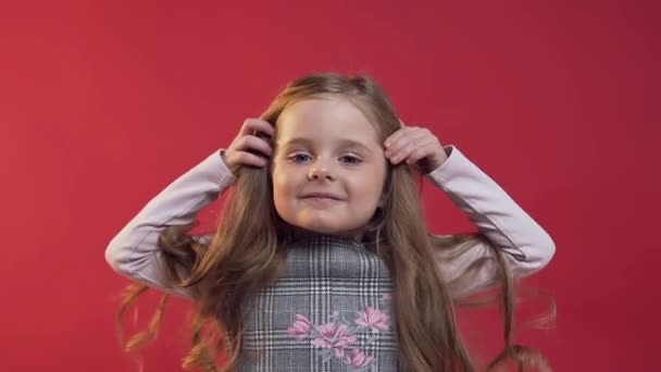 Закрывается симпатичная 5-летняя девочка с длинными кудрявыми волосами, позирующая на камеру на фоне красной стены — стоковое видео