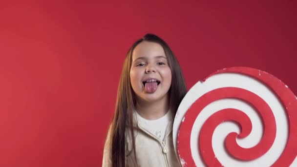 Close-up van grappig glimlachend tiener meisje met bretels die tong tonen, plastic Lollipop in de hand houden op de rode achtergrond — Stockvideo