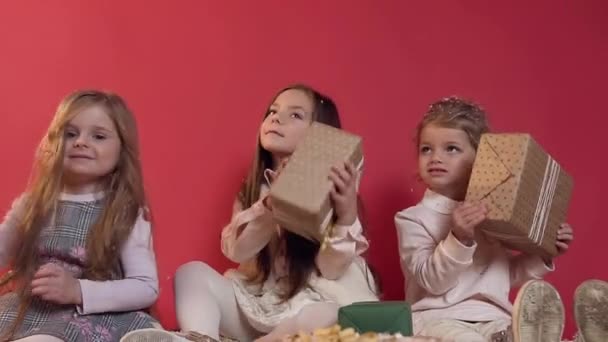 Tři roztomilí šťastní modelky, které sedí na podlaze na červeném pozadí a hrají s dobře zabalenými dárky — Stock video
