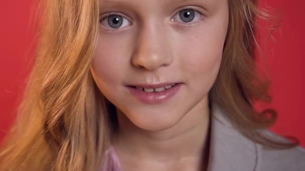 Очаровательная улыбающаяся девушка-подросток с светлыми волосами и голубыми глазами, смотрящая в камеру на красном фоне — стоковое видео