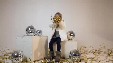 Disko topları yakınında oturan ve beyaz arka plan üzerinde kameraya altın konfeti üfleme moda giysileri oldukça neşeli genç model yavaş hareket