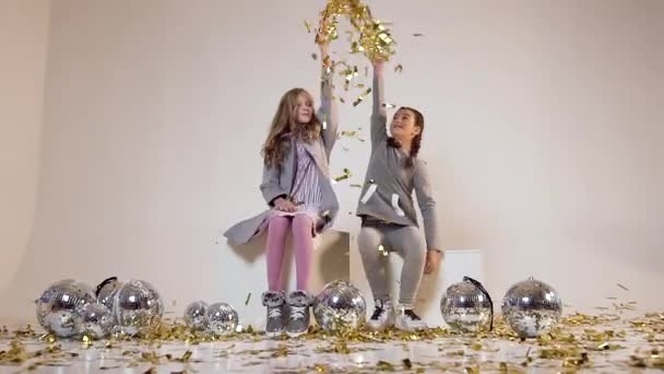 Movimento lento de lindas e alegres duas meninas adolescentes sentadas perto de bolas de discoteca e jogando confetes dourados sobre suas cabeças no fundo branco — Vídeo de Stock