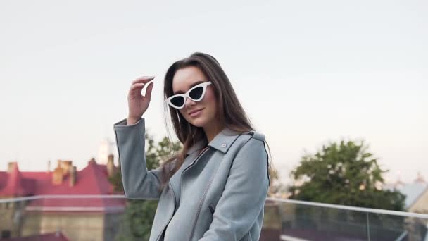 Belle jeune femme souriante dans des vêtements à la mode assis sur le vélo et regardant la caméra dans des lunettes de mode sur la terrasse — Video