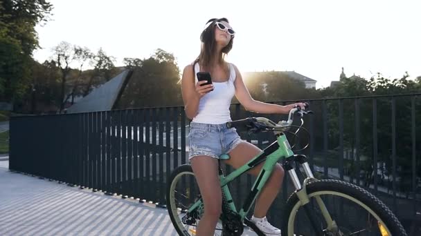Горячая улыбчивая молодая женщина в стильных очках сидит на велосипеде и слушает музыку в наушниках на фоне города — стоковое видео