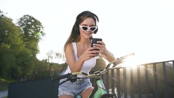 Close up de sorrir jovem mulher fofa em fones de ouvido sentado na bicicleta e ouvir música — Vídeo de Stock