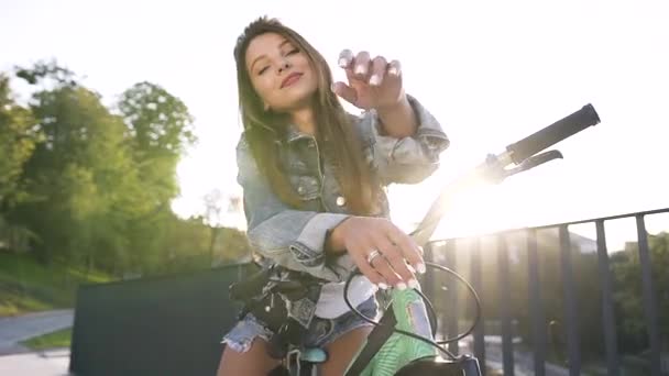 Menina sexy exuberante impressionante em roupas da moda sentado na bicicleta e olhando para a câmera com sorriso bonito no fundo do pôr do sol — Vídeo de Stock