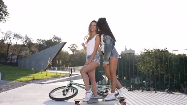 Two cuddy jovial sexy girls in fashion clothes standing by one leg on skateboard with raised hands and posing on camera with cute smiles on city background — Stock Video