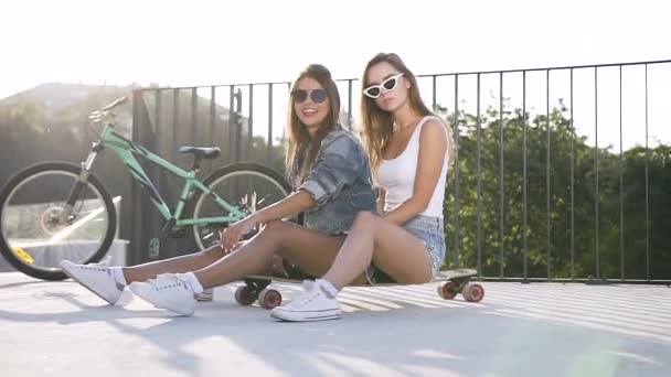 Atraente feliz meninas sexy sentados no skate e posando na câmera com sorrisos agradáveis na paisagem da cidade — Vídeo de Stock