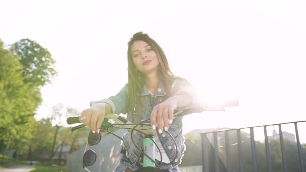 Mujer sexy alegre de moda con el pelo largo marrón sentado en bicicleta y mirando a la cámara, lleva gafas de sol en el día soleado — Vídeos de Stock