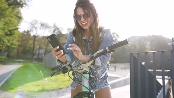 自転車に座って、街の背景に彼女の携帯電話を使用してジーンズの服を着て気楽な笑顔若い女性 — ストック動画