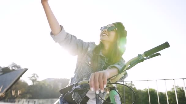 Menina alegre lindo em óculos de sol modish e roupas elegantes sentado na bicicleta e fazendo selfie engraçado sob as luzes do sol — Vídeo de Stock