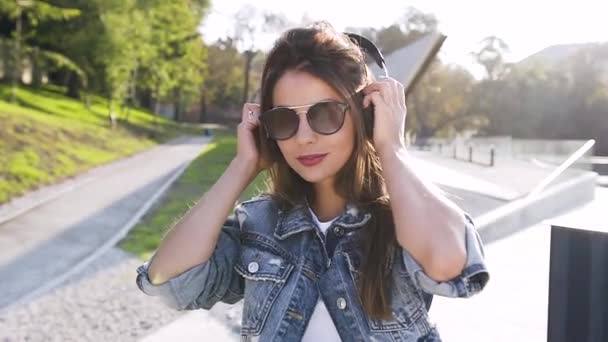 Bonita mujer de 30 años con gafas de sol con auriculares y música en el fondo del paisaje de la ciudad — Vídeo de stock