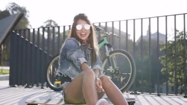 Camları ve kot pantolon giysileri ile koyu saçlı çarpıcı gülümseyen kadın yakın kaykay üzerinde oturan ve yeşil şehir sokakta kamera bakarak