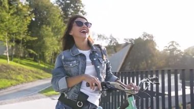 Şık güneş gözlüğü muhteşem gülümseyen kız, güneşli bir günde çit yakınında bisiklet ile gidiyor trendy giysiler giymiş