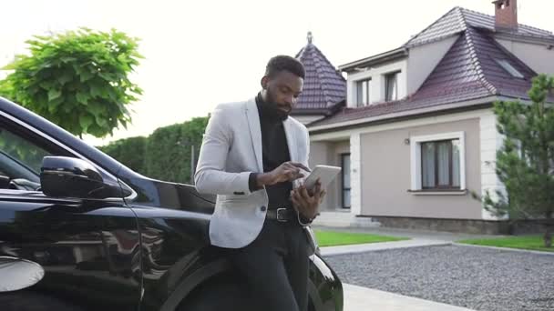 Knappe jonge Afro-Amerikaan met behulp van sms-app op computer tablet terwijl hij op parkeren in de buurt van luxe zwarte auto in gezellige stad. — Stockvideo