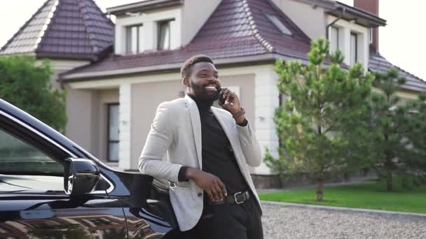 Guapo joven afroamericano responde a una importante llamada telefónica mientras está de pie en el aparcamiento cerca de coche negro de lujo en la acogedora ciudad . — Vídeos de Stock