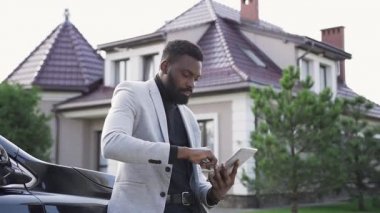 Başarılı genç Afrikalı Amerikalı adam, dokunmatik ekran dijital tabletine tıklayarak tablet bilgisayar aygıtı kullanırken rahat bir kasabada siyah lüks arabanın yanında duruyor.