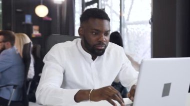 Çekici sakallı Afro-Amerikan ofis çalışanı bilgisayarın başında çalışıyor ve kameraya sevimli bir gülümsemeyle poz veriyor.