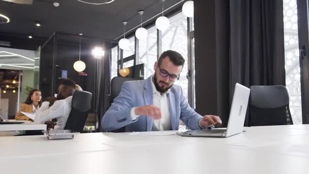 彼のコンピュータ、異なる財務報告書を使用し、企業の同僚の背景に電話で話す目的のハンサムなオフィスの労働者 — ストック動画