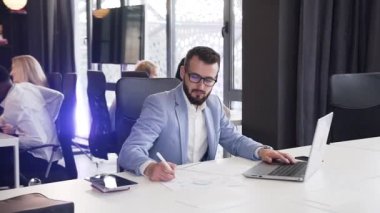 İyi görünümlü sakallı ofis yöneticisi iş belgeleriyle çalışıyor ve ofis odasındaki bilgisayarlara veri girişi yapıyor.