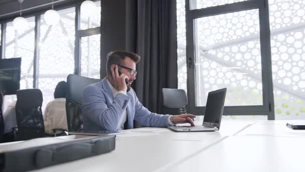Серьезный высококвалифицированный бизнесмен обсуждает компьютерный проект по телефону с собеседником, сидящим на рабочем месте в офисе — стоковое видео