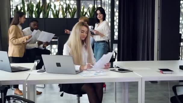 Bonita jovem empresária cuidadosa com cabelo loiro longo sentado no local de trabalho, olhando para os relatórios e usando o computador na sala de escritório — Vídeo de Stock