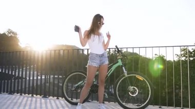 Kot şortlu ve tişörtlü çekici kız elinde akıllı telefon, bisiklet sürdükten sonra sokak ortasında dans ediyor. Dışarıda.