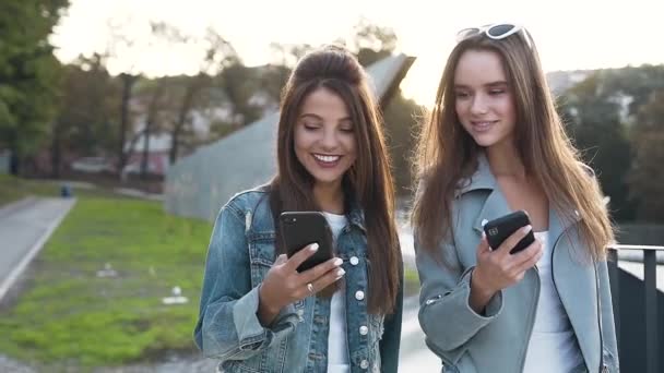 Portrait of two young attractive female friends with long hair using smartphone while walking laughing and watching something in app on the smartphone screen. — Stock Video