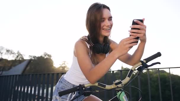 Πορτρέτο της ελκυστικό χαμογελαστό κορίτσι με μακριά μαλλιά κάθεται στο ποδήλατο και κάτι βλέποντας σε εφαρμογή smartphone το καλοκαίρι στην πόλη — Αρχείο Βίντεο