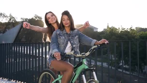 Alegre meninas amigas caucasianas em roupas da moda com uma bicicleta andando na cidade ao ar livre, abraçando e sorrindo alegremente à noite de verão . — Vídeo de Stock