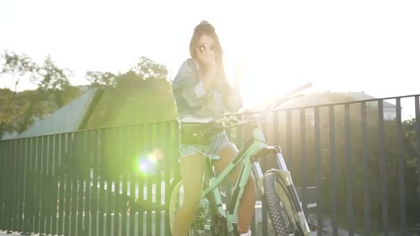 Attraktiv urban flicka i snygga kläder, solglasögon med vackra långa hår dansar i staden när du står med cykel, har attraktiva leende, klädd i jacka och jeans shorts på sommaren. — Stockvideo