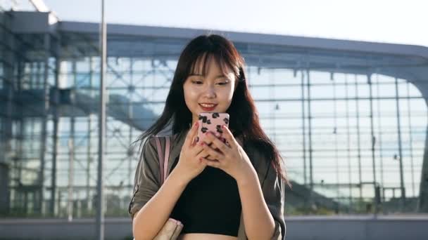 Nära bild av Jocund glad 25-årig asiatisk dam med långt hår och snygga kläder som använder sin smartphone nära modern flygplats byggnad — Stockvideo