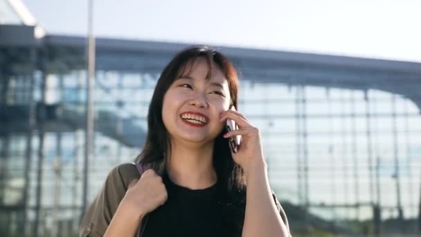 Ganska glad ung asiatisk kvinna med mörkt hår talar i telefon och uppriktigt leende nära flygplatsen byggnad på dagen — Stockvideo