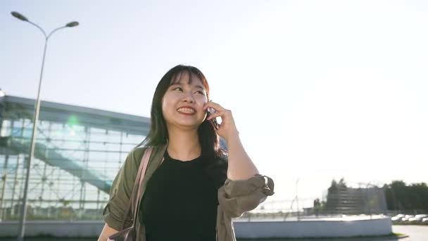 Bonito jinete de 25 años morena asiática hablando en el móvil cerca del edificio del aeropuerto en el día — Vídeo de stock