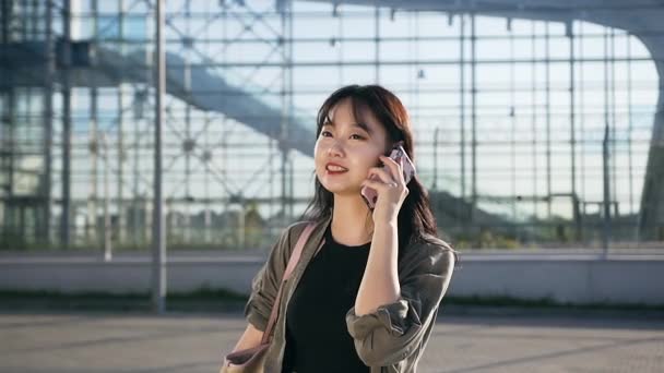 Vonzó vidám ázsiai fiatal nő beszél okostelefonon a háttérben a repülőtér építése — Stock videók