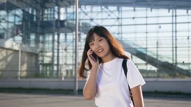 Aangename glimlach jong aziatisch meisje met donker lang haar praten op mobiele in de buurt van de luchthaven in de dag — Stockvideo