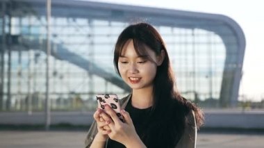 Güzel gülümseyen Asyalı kadın havaalanı binasının yanında yürüyor ve cep telefonunu kullanıyor.
