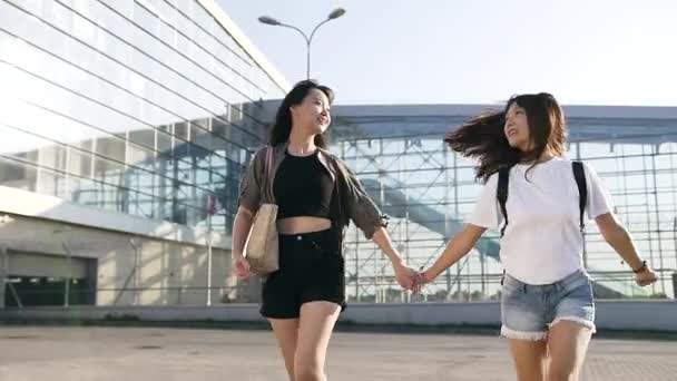 Atractiva imagen de las niñas asiáticas sonrientes felices en ropa de moda cogidas de la mano y divirtiéndose juntos cerca del edificio del aeropuerto en el día — Vídeo de stock