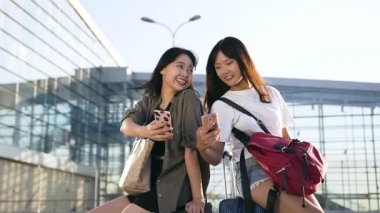 Bavullarının üzerinde oturan ve büyük havaalanı terminalinin yanındaki akıllı telefonları kullanan iki çekici Asyalı kadının ön görüntüsü.