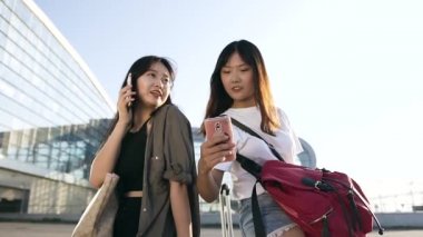 İyi görünümlü 25 'li yaşlarda Asyalı kız arkadaşlar bavullarının üzerinde oturuyorlar ve modern havaalanı terminalinin yanında akıllı telefonlar kullanıyorlar.