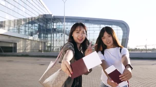 Close view van opgetogen goed uitziende Aziatische vrouwen met paspoorten en tickets in hun handen die kijken naar camera met een vrolijke glimlach in de buurt van de terminal van de luchthaven — Stockvideo