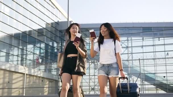 Крупним планом усміхнені щасливі милі азіатські дівчата в модному молодіжному одязі зі своїми валізами та документами, які ходять біля великої будівлі аеропорту — стокове відео