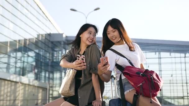 Вид спереди двух очаровательных эксцентричных женщин, сидящих на чемоданах и пользующихся смартфонами возле большого терминала аэропорта — стоковое видео