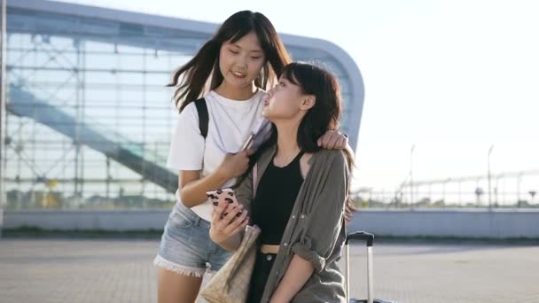 25-летняя девушка с длинными волосами смотрит в телефон и улыбается возле современного здания аэропорта — стоковое видео