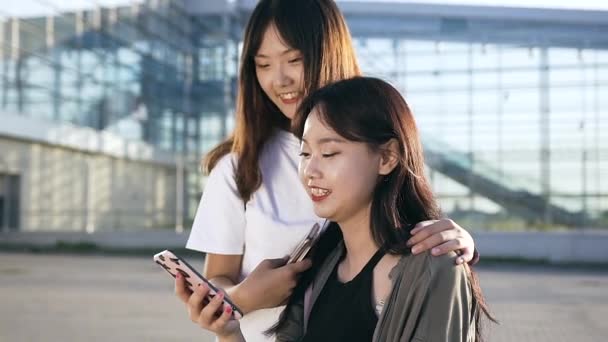 Dvě krásné bujné mladé asijské dívky se usmívají z revize legrační fotografie na telefonu v blízkosti moderní budovy — Stock video